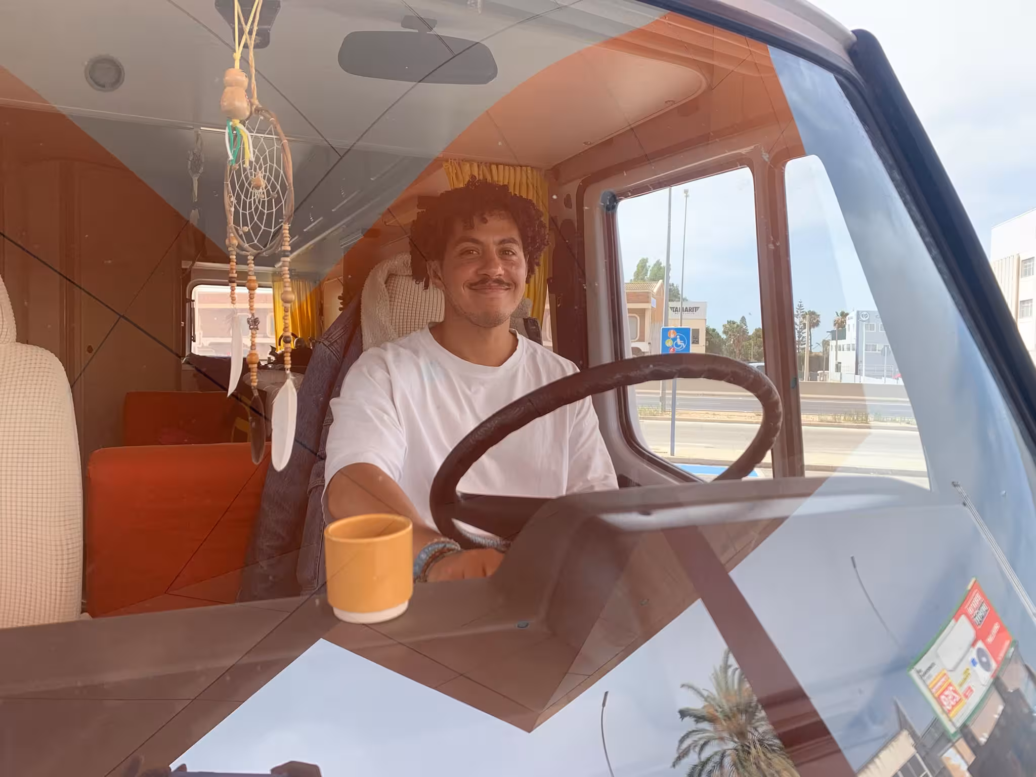 Ahmed smiling behind the steering wheel inside a van with beige and orange interior, a yellow curtain behind him, and a coffee mug on the dashboard.